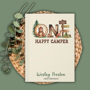 Puede incluir: Un libro de color crema con un tema de bosque. La portada presenta la palabra "ONE" en letras marrones con ilustraciones de un oso, un zorro y una tienda de campa&ntilde;a. El texto "Happy Camper" est&aacute; debajo de la palabra "ONE". La parte inferior de la portada dice "Wesley Preston Primer Cumplea&ntilde;os".