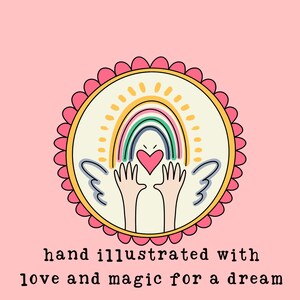 May include: A hand-drawn illustration of a rainbow with a heart in the center, surrounded by two hands with wings. The illustration is in a circle with a pink and yellow border. The text "hand illustrated with love and magic for a dream" is below the circle.