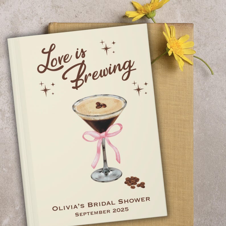 May include: A cream-colored bridal shower guest book with the words "Love is Brewing" in brown script. The cover features a watercolor illustration of a coffee martini with a pink ribbon and coffee beans. The text "Olivia's Bridal Shower September 2025" is printed at the bottom.