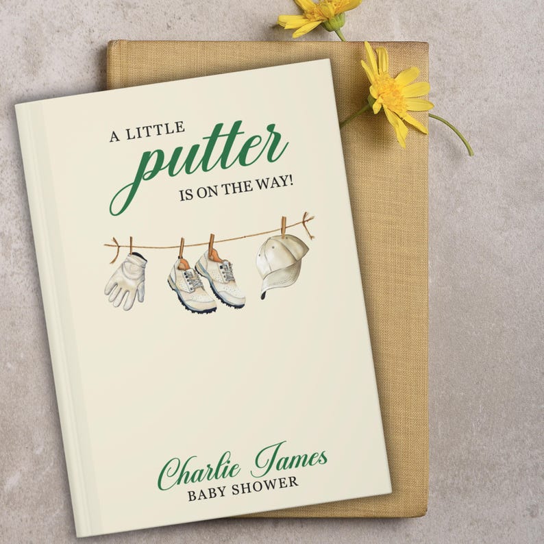 May include: A cream-coloured baby shower book with the text "A LITTLE putter IS ON THE WAY!" in green script. The cover features a watercolour illustration of golf-related items hanging on a washing line. The name "Charlie James" and "BABY SHOWER" are printed below.