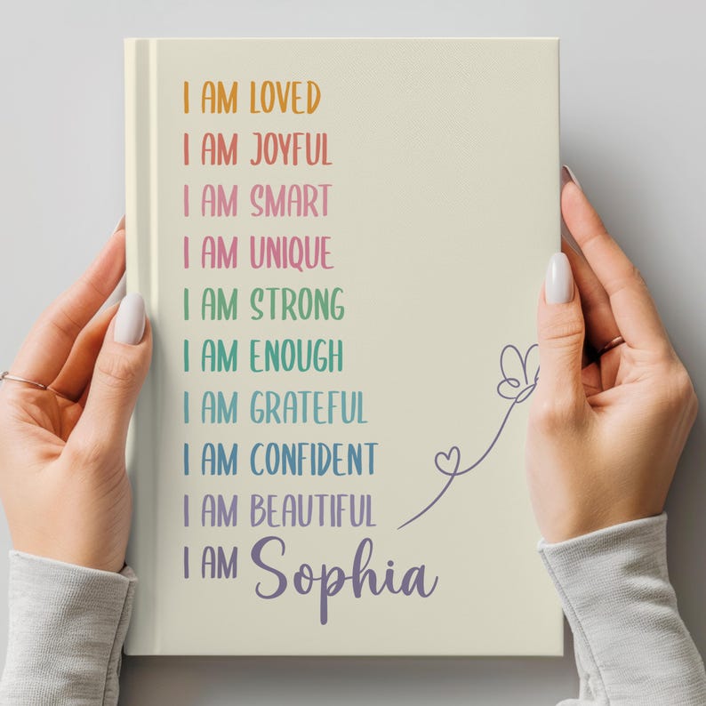 Pu&ograve; includere: Un diario color crema con le parole "I AM LOVED, I AM JOYFUL, I AM SMART, I AM UNIQUE, I AM STRONG, I AM ENOUGH, I AM GRATEFUL, I AM CONFIDENT, I AM BEAUTIFUL, I AM Sophia" in vari colori. Una farfalla e un cuore sono disegnati sul lato destro.