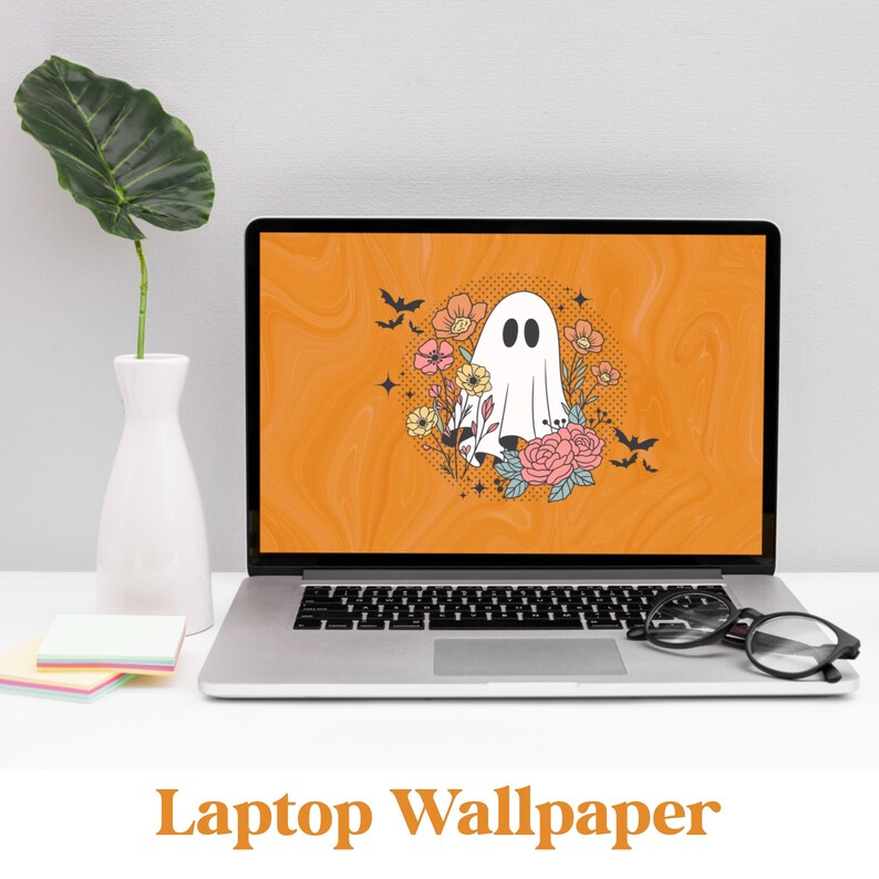 May include: A laptop computer with a Halloween-themed wallpaper featuring a white ghost with a floral wreath around it on an orange background. The text "Laptop Wallpaper" is displayed below the laptop.