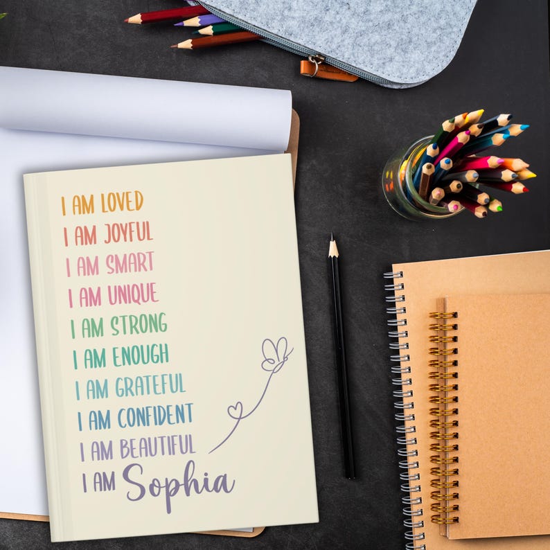 Pu&ograve; includere: Un quaderno color crema con il testo "I AM LOVED, I AM JOYFUL, I AM SMART, I AM UNIQUE, I AM STRONG, I AM ENOUGH, I AM GRATEFUL, I AM CONFIDENT, I AM BEAUTIFUL, I AM Sophia". Una matita nera, matite colorate e altri quaderni sono visibili.