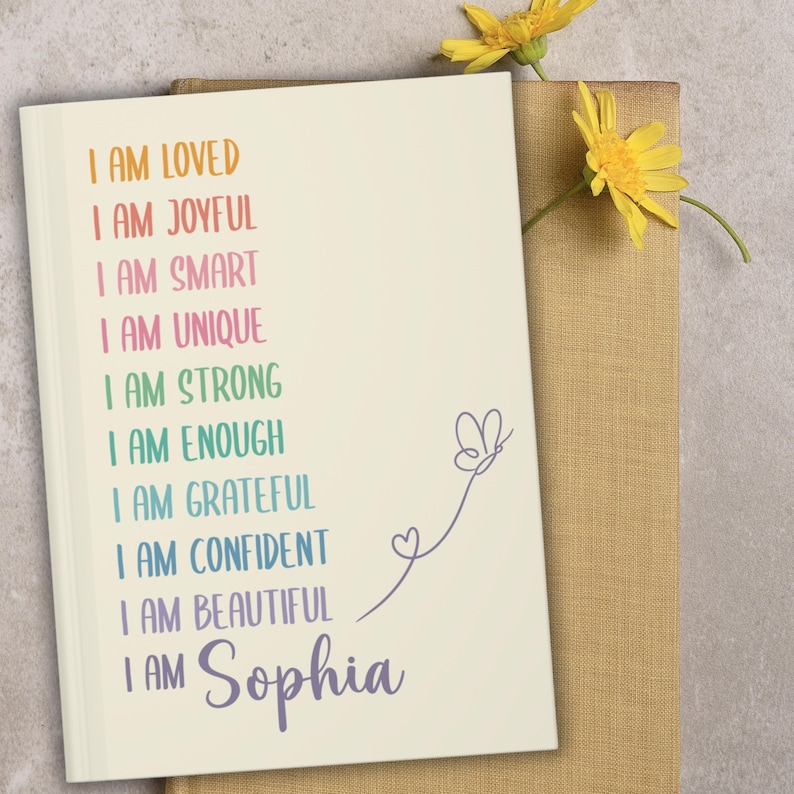 Pu&ograve; includere: Un quaderno color crema con testo colorato che recita "I AM LOVED, I AM JOYFUL, I AM SMART, I AM UNIQUE, I AM STRONG, I AM ENOUGH, I AM GRATEFUL, I AM CONFIDENT, I AM BEAUTIFUL, I AM Sophia." Una farfalla e un cuore sono disegnati sul lato destro. Fiori gialli sullo sfondo.