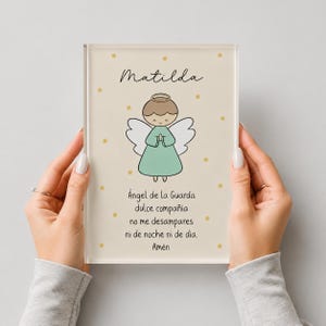 May include: A rectangular acrylic plaque featuring a cartoon angel with a halo and light green robe. The name Matilda is printed at the top, and the Spanish prayer "Ángel de la Guarda" is printed below the angel, surrounded by small gold stars.