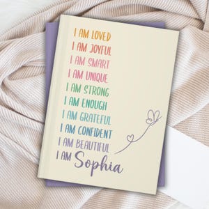 Pu&ograve; includere: Un quaderno color crema con le parole "I AM LOVED, I AM JOYFUL, I AM SMART, I AM UNIQUE, I AM STRONG, I AM ENOUGH, I AM GRATEFUL, I AM CONFIDENT, I AM BEAUTIFUL, I AM Sophia" in colori arcobaleno. Una farfalla e un cuore sono disegnati sul lato destro.