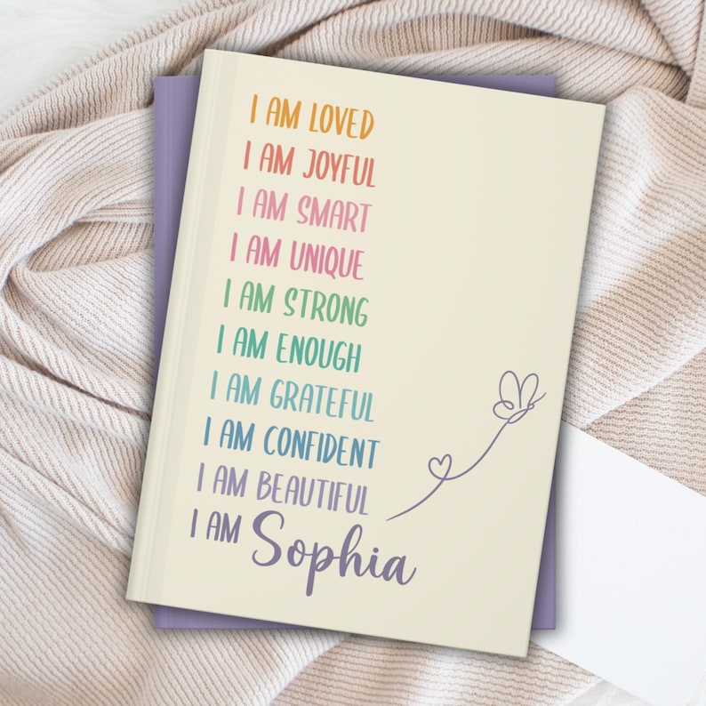 Pu&ograve; includere: Un quaderno color crema con le parole "I AM LOVED, I AM JOYFUL, I AM SMART, I AM UNIQUE, I AM STRONG, I AM ENOUGH, I AM GRATEFUL, I AM CONFIDENT, I AM BEAUTIFUL, I AM Sophia" in colori arcobaleno. Una farfalla e un cuore sono disegnati sul lato destro.
