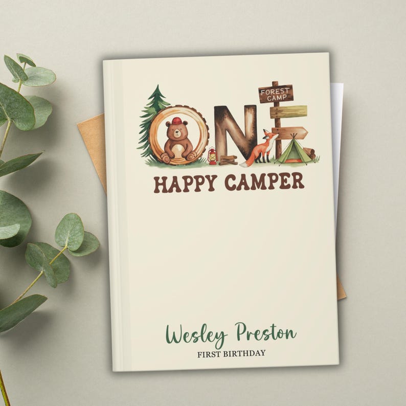 Puede incluir: Un diario de color crema con un tema de bosque. La portada presenta la palabra "ONE" con un oso, un zorro y equipo de campamento. El texto "Happy Camper" est&aacute; debajo de la palabra "ONE". La parte inferior de la portada dice "Wesley Preston Primer Cumplea&ntilde;os".