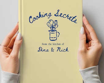 May include: A light yellow cookbook titled "Cooking Secrets" with blue lettering and a drawing of kitchen utensils. The text "from the kitchen of Dina & Nick" is also in blue. The book is held by two hands.