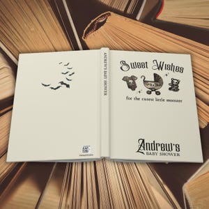 May include: An open book with a cream-colored cover featuring a bat silhouette and the text "Sweet Wishes for the cutest little monster." The book is for Andrew's baby shower. The book is resting on a background of open books.
