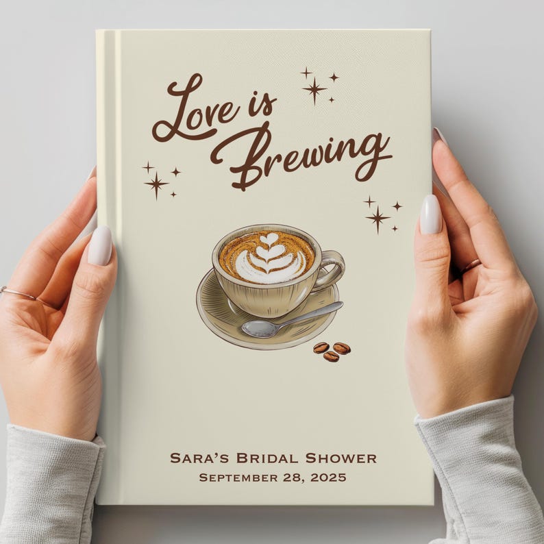 May include: A cream-colored book cover with the words "Love is Brewing" in brown script, along with a coffee cup illustration. The text "Sara's Bridal Shower, September 28, 2025" is printed at the bottom. The book is held by two hands.