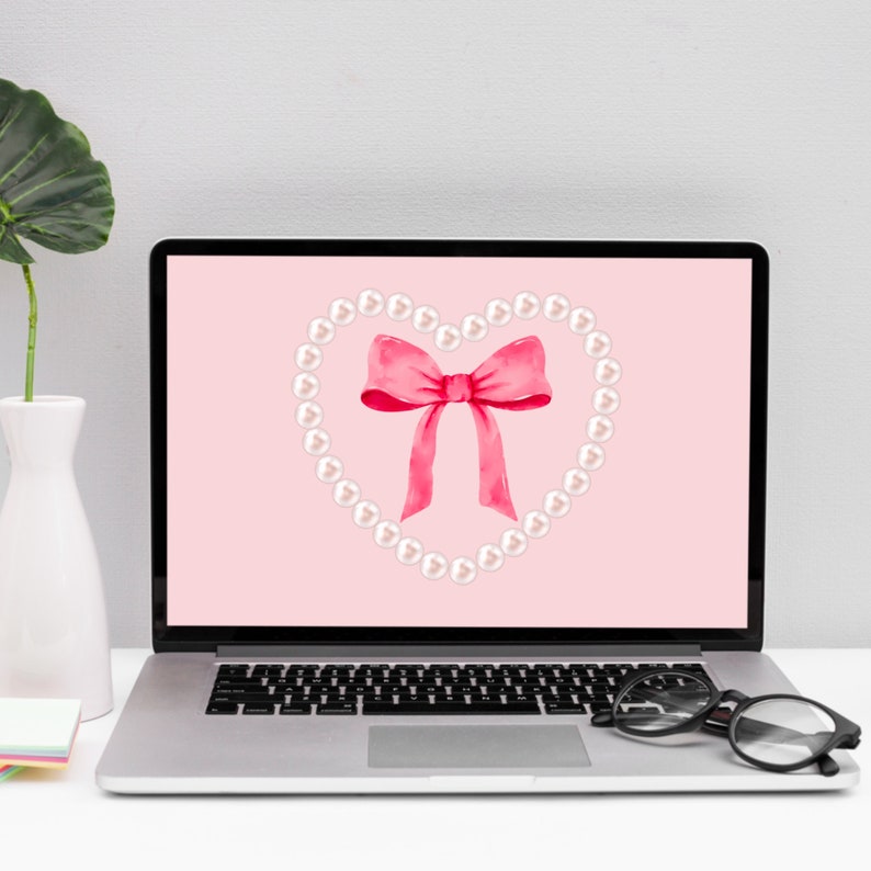 May include: A laptop computer with a pink background and a white pearl heart shape with a pink bow in the center. The laptop is on a white surface.