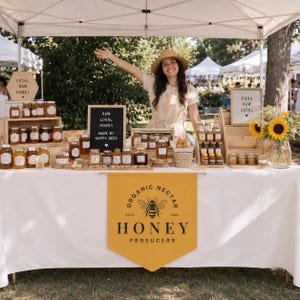 May include: A market stall featuring numerous glass jars of honey arranged on wooden crates and shelves. A yellow banner displays the text Organic Nectar Honey Producers Estd 2020 with a bee graphic. Letter boards read Local Raw Honey and Pure Raw Local.