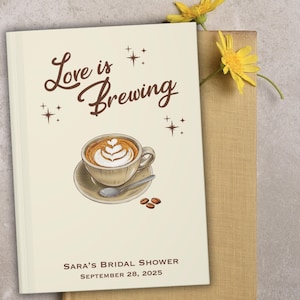 May include: A cream-colored bridal shower guest book with the text "Love is Brewing" and a coffee cup illustration. The book includes the text "Sara's Bridal Shower, September 28, 2025". Yellow flowers are placed on top of a tan book.