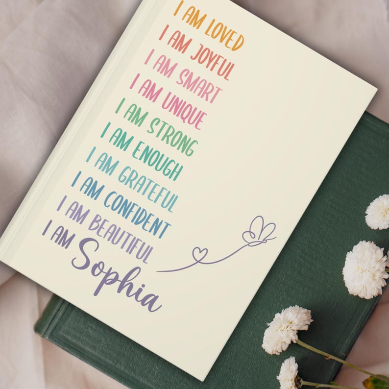 Pu&ograve; includere: Un diario color crema con le parole "I AM LOVED, I AM JOYFUL, I AM SMART, I AM UNIQUE, I AM STRONG, I AM ENOUGH, I AM GRATEFUL, I AM CONFIDENT, I AM BEAUTIFUL, I AM Sophia" in testo colorato. Un disegno di farfalla e cuore &egrave; sulla copertina.
