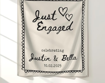 May include: A cream-colored fabric banner with black text and a decorative border. The banner reads "Just Engaged" with two hearts, followed by "celebrating Justin & Bella" and the date 10.02.2025. The banner is hanging on a white wall.