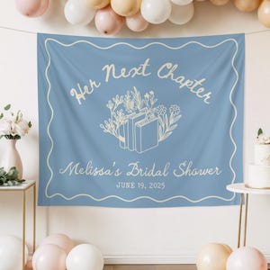 Op de afbeelding: Een lichtblauwe banner met de tekst "Her Next Chapter" en "Melissa's Bridal Shower" in een scriptlettertype. De banner heeft een bloemen- en boekontwerp. De datum "June 19, 2025" is ook opgenomen. Een witte geschulpte rand omlijst de tekst.
