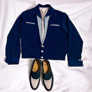 Könnte beinhalten: Eine marineblaue Bomberjacke mit einem weißen und blauen Karomuster auf der Vorderseite. Die Jacke hat einen Reißverschluss und einen gerippten Saum. Zwei dunkelblaue Schuhe mit weißen Verzierungen sind ebenfalls abgebildet.