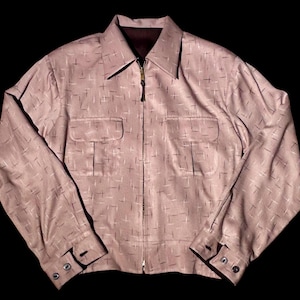 May include: A light mauve long-sleeve jacket featuring a repeating pattern of small dark cross-hatch marks. The garment includes a pointed collar, a full-length front zipper with a dark pull, two chest pockets with flaps, and buttoned cuffs.