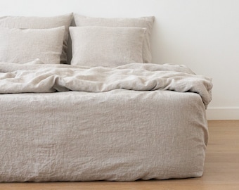 May include: A beige linen bed with a white wall in the background. The bed is made with a linen duvet and two linen pillows.