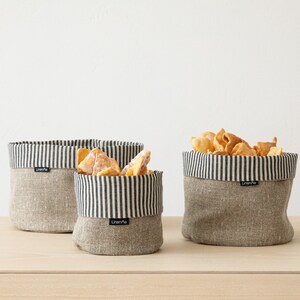May include: Three gray linen storage baskets with black and white striped fabric lining. The baskets are on a light wood surface. The basket on the right contains a pile of fried dough.