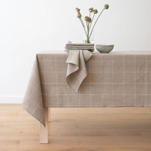 Op de afbeelding: Een bruine linnen tafelkleed met een wit geruit patroon. Het tafelkleed is gedrapeerd over een houten tafel met een vaas met gedroogde bloemen en een kom erop.