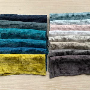 Puede incluir: Una colección de paños de cocina plegados con tejido de gofre en varios colores, incluyendo azul marino, turquesa, amarillo y gris. Las toallas están cuidadosamente apiladas sobre una superficie de madera clara, mostrando su diseño texturizado y variaciones de color. Cada toalla mide aproximadamente 30 cm de ancho.