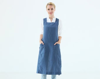 May include: A woman wearing a long, blue linen apron with two large pockets. The apron has a square neckline and is tied at the waist. She is standing in front of a white background.