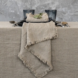 Puede incluir: Una mesa de lino con una paleta de colores neutros. La imagen presenta un mantel de lino beige, servilletas a juego con bordes deshilachados, platos negros y vasos azul oscuro. La cubertería y una ramita de romero completan el elegante arreglo.