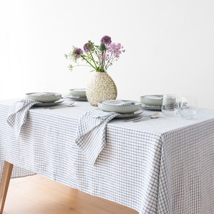Può includere: Una tovaglia bianca con un motivo a quadri blu, apparecchiata per una cena con quattro coperti. Ogni coperto comprende un tovagliolo bianco, un piatto e posate. Un vaso con fiori viola si trova al centro del tavolo.