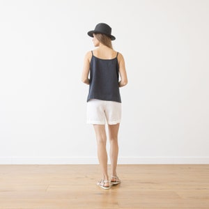 May include: A woman wearing a dark gray linen tank top and white linen shorts. She is wearing a black fedora hat and brown sandals.