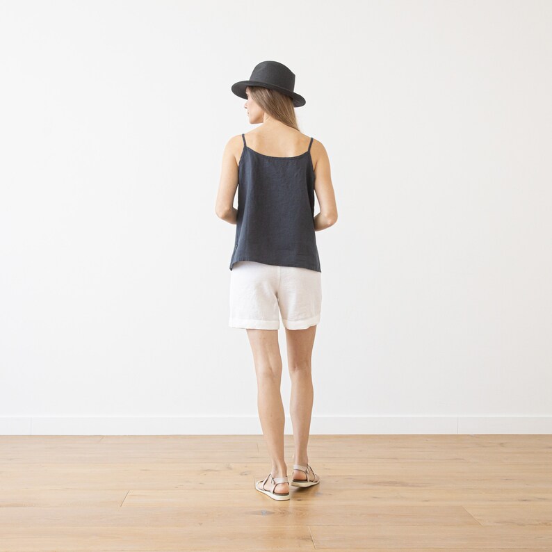 May include: A woman wearing a dark gray linen tank top and white linen shorts. She is wearing a black fedora hat and brown sandals.