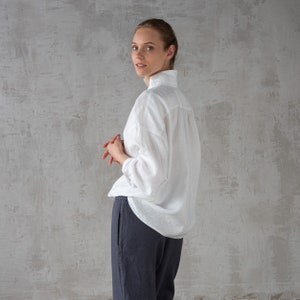 K&ouml;nnte beinhalten: Ein wei&szlig;es Leinenhemd mit Stehkragen und langen &Auml;rmeln. Das Hemd ist locker geschnitten und hat eine lockere Silhouette. Das Model tr&auml;gt das Hemd zu einer dunkelgrauen Hose.