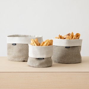 May include: Three linen storage baskets in varying sizes. The baskets are a natural beige color with a striped, off-white and beige, fabric lining. The baskets are filled with snacks. The brand name "LinenMe" is printed on a small tag on each basket.