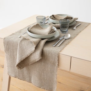 Può includere: Una tavola apparecchiata con un runner in lino beige con bordo sfilacciato. L'allestimento comprende piatti, ciotole, tovaglioli, posate e due bicchieri d'acqua. Lo stile è minimalista e naturale, adatto per una sala da pranzo.