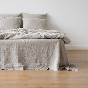 May include: A beige linen bed with a white headboard and a beige linen bed skirt. The bed is made with a beige linen duvet cover and two beige linen pillows.