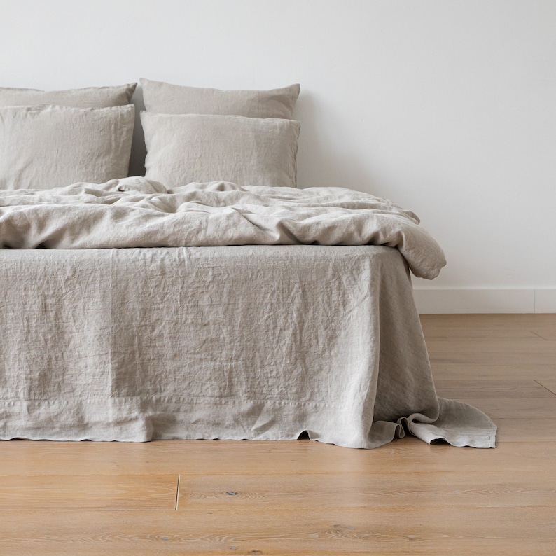May include: A beige linen bed with a white headboard and a beige linen bed skirt. The bed is made with a beige linen duvet cover and two beige linen pillows.
