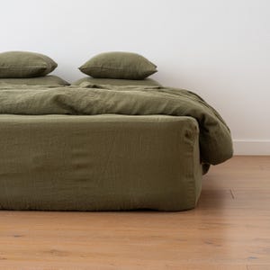 May include: An olive green linen bedding set, comprising a fitted sheet, duvet cover, and pillowcases. The bed is made up with pillows stacked on top. The fabric has a slightly wrinkled texture, suggesting a relaxed, comfortable aesthetic.