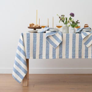 May include: A blue and white striped linen tablecloth on a wooden table, set for a meal. Napkins and candles are arranged on the table. The tablecloth has vertical stripes, creating a fresh and inviting atmosphere.
