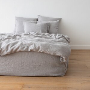 May include: A gray linen duvet cover and pillowcases are arranged on a bed with a gray linen bed skirt. The bed is made with a simple, minimalist style.