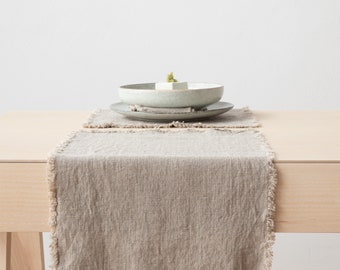 Puede incluir: Un camino de mesa de lino beige con un borde deshilachado, colocado sobre una mesa de madera clara. Se ve un mantel individual a juego y dos platos de cerámica con comida, creando un ambiente de comedor minimalista.