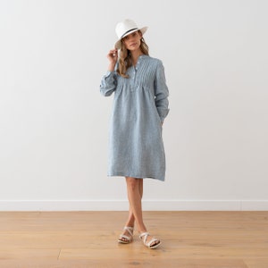 May include: A light blue linen dress with a white button-up collar and a white hat. The dress has a pleated detail at the chest and a relaxed fit.