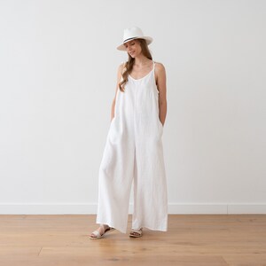 May include: A white linen jumpsuit with thin straps and wide legs. The model is wearing a white hat and sandals.
