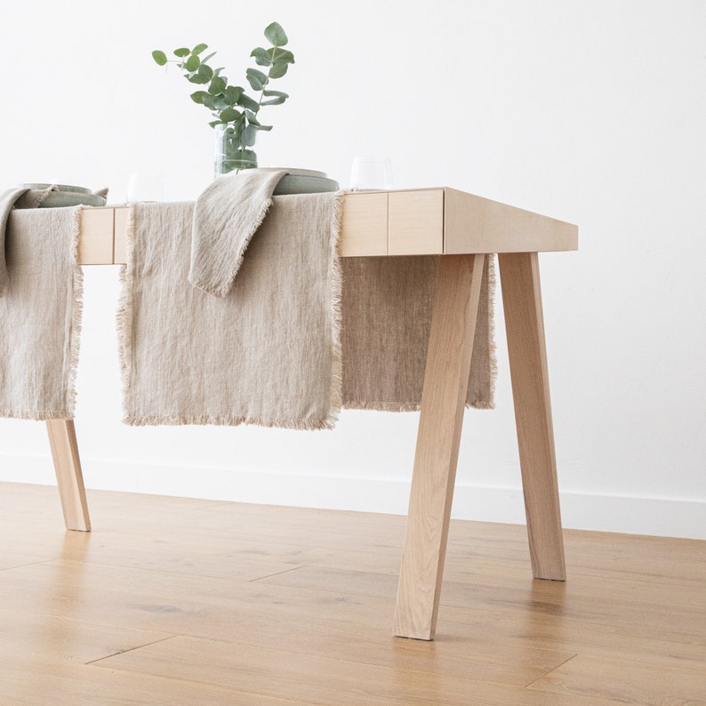 May include: A light wood table with a natural finish and four legs. Two beige linen table runners are draped over the table.