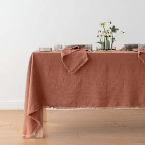 Puede incluir: Un mantel de lino color terracota con borde deshilachado cubre una mesa de madera. La mesa está puesta con servilletas, platos y cristalería a juego. Un pequeño jarrón de flores está en el centro.