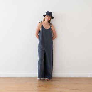 May include: A woman wearing a dark blue linen jumpsuit with thin straps and a black fedora hat. She is standing in front of a white wall.