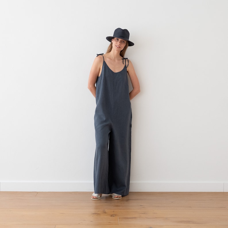 May include: A woman wearing a dark blue linen jumpsuit with thin straps and a black fedora hat. She is standing in front of a white wall.