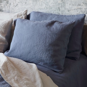 Könnte beinhalten: Nahaufnahme eines Bettes mit blauer und cremefarbener Leinenbettwäsche. Zwei große, quadratische blaue Kissen sind der Mittelpunkt, mit einer zerknitterten Textur. Ein weißes Laken und ein Kissen sind im Hintergrund zu sehen.