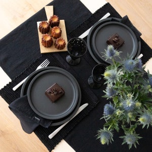 May include: Two black plates with a black and white dessert on each plate. The plates are on a black tablecloth with a wooden cutting board with small pastries on it. A bouquet of blue flowers is in the center of the table.