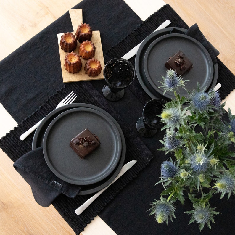 May include: Two black plates with a black and white dessert on each plate. The plates are on a black tablecloth with a wooden cutting board with small pastries on it. A bouquet of blue flowers is in the center of the table.